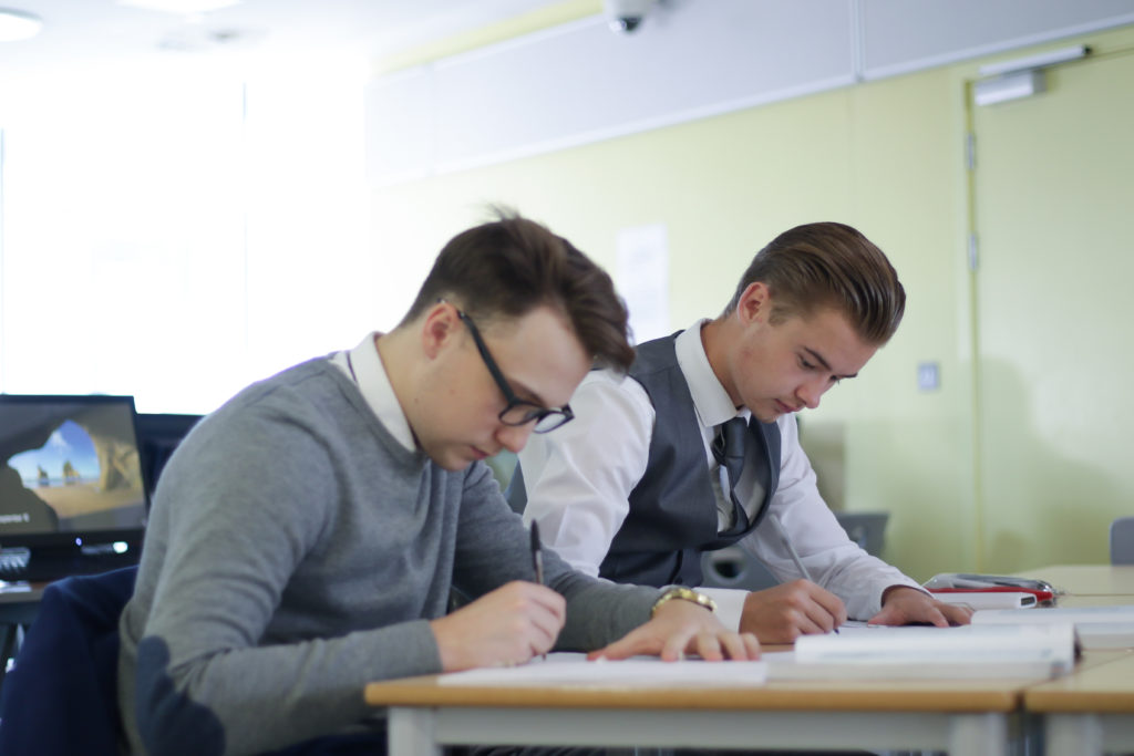 Two male Post-16 Leigh Academy Longfield students sit together whilst writing on paper with pens.