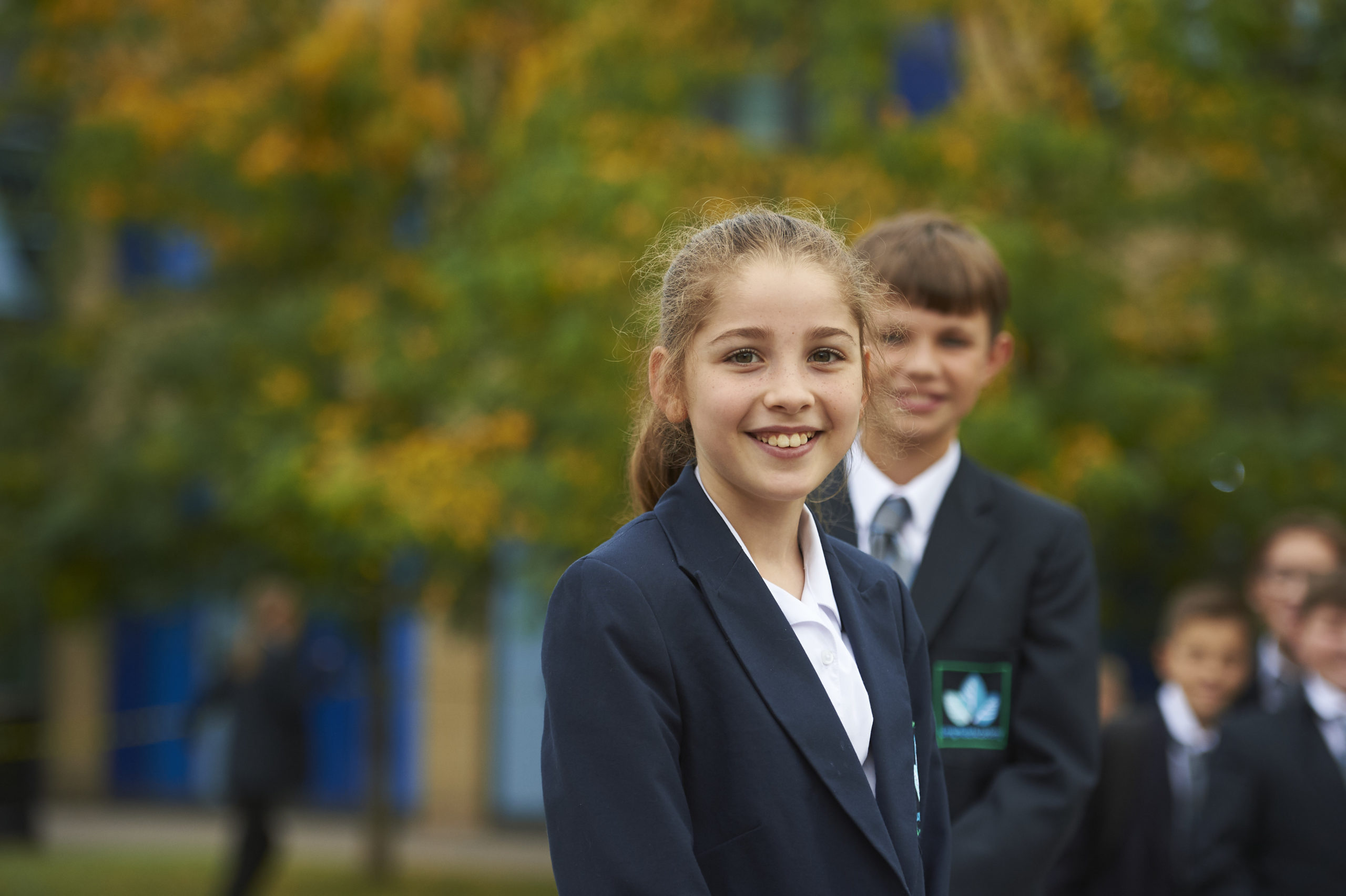 Two Leigh Academy Longfield students are pictured dressed in their academy uniform and smiling for the camera. A small group of students can be seen in the background behind them.