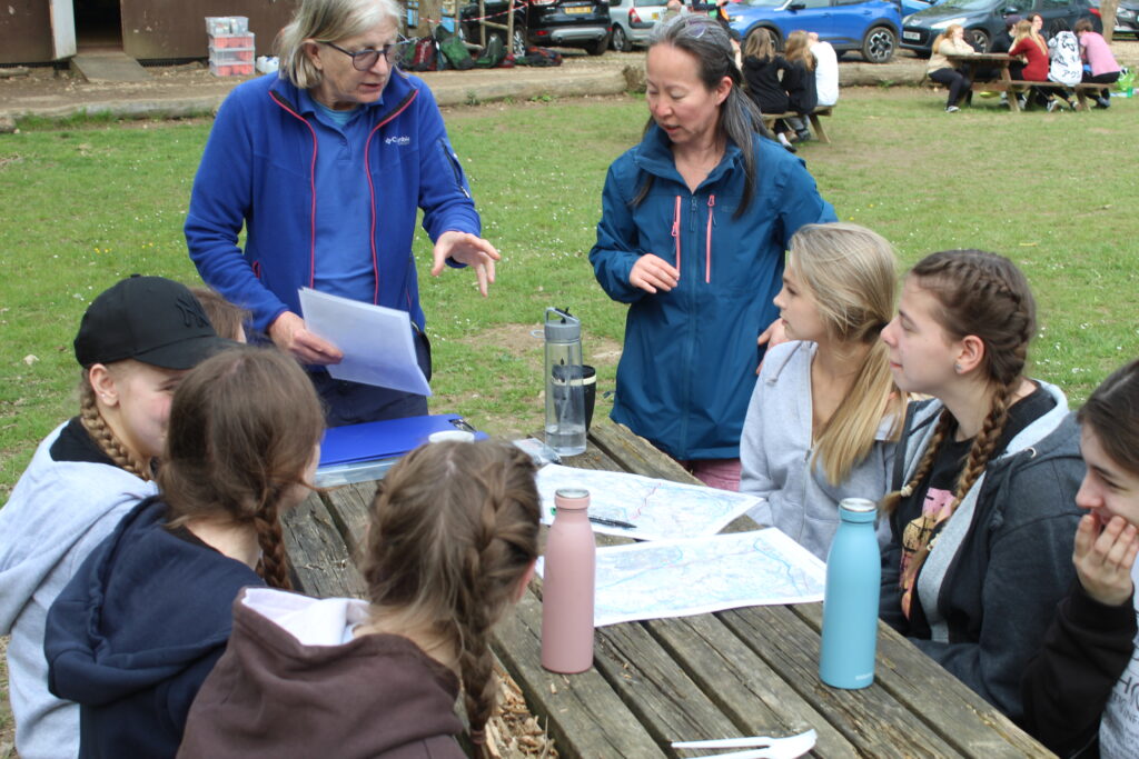 A group of students are shown sitting at a wooden table outdoors and listening to the instructions of a member of staff.
