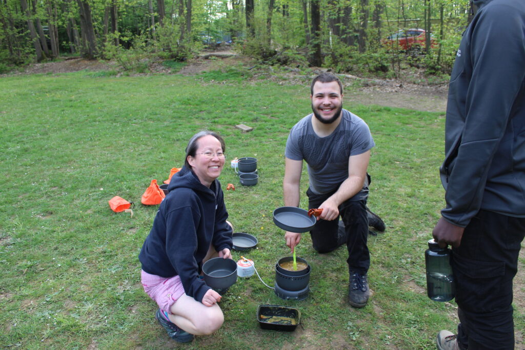 A student and female member of staff are shown sitting together on the grass, smiling for the camera whilst cooking a meal for the group,