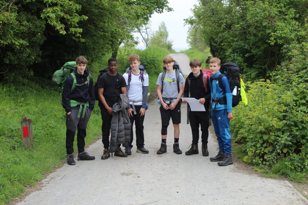 Six male students are pictured smiling for the camera on a paved footpath in the woods, wearing their backpacks on their backs.