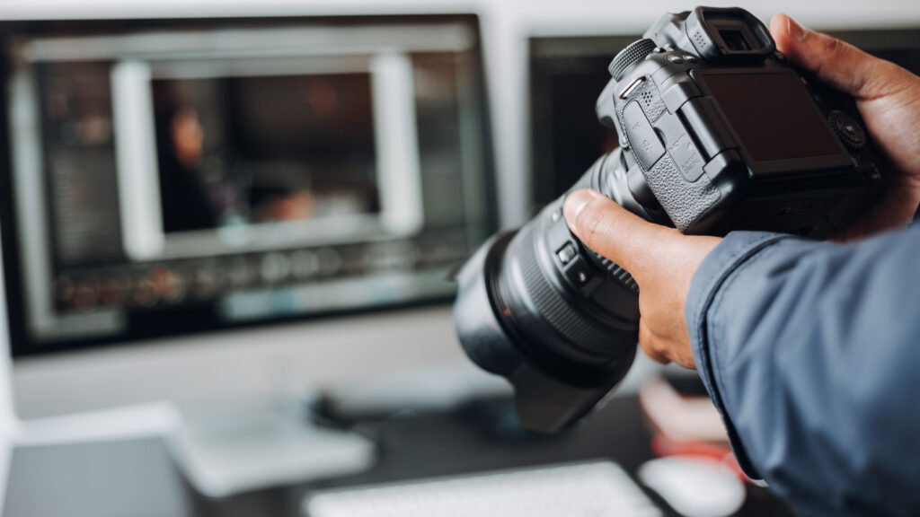 A pair of hands are seen holding a professional camera with a laptop seen sat on a desk in the background.