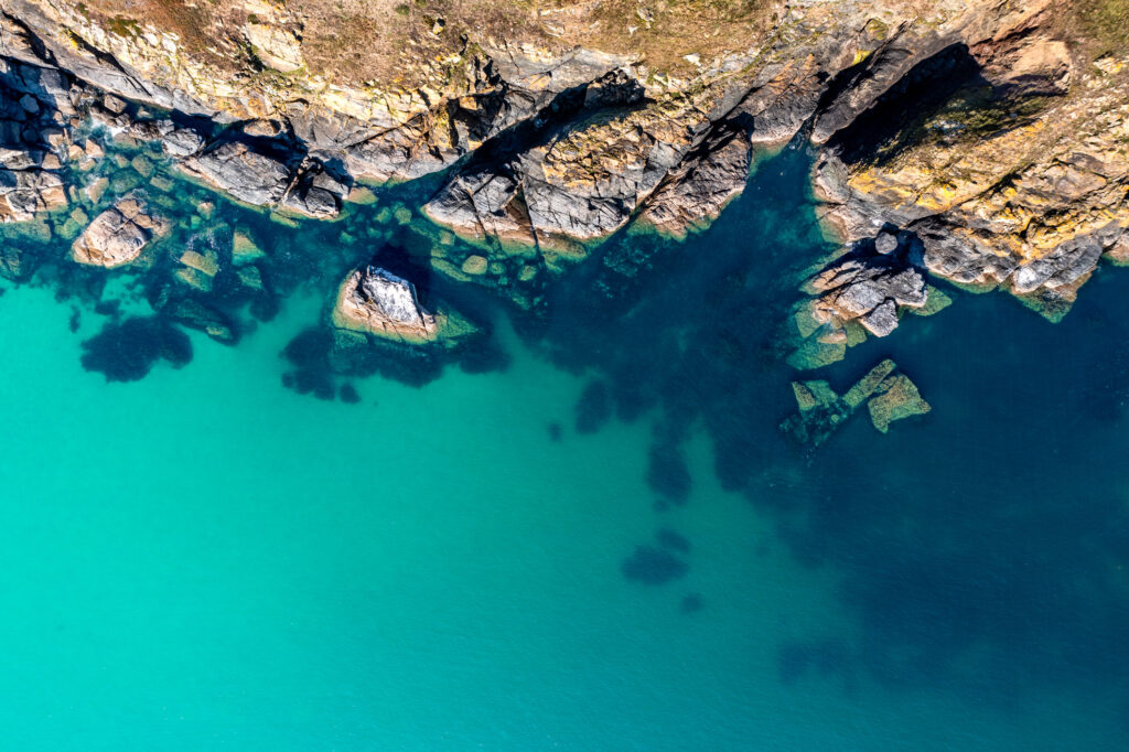 Aerial view directly above rugged rocky outcrop of land jutting into an emerald green ocean with sand sea bed in Cornwall, UK with copy space