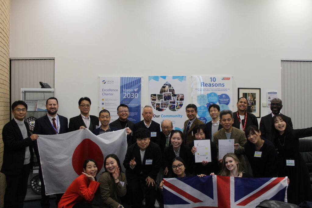 A group of people, appearing to be adults and young adults, posing together in an indoor setting with a Japanese flag and a Union Jack flag.